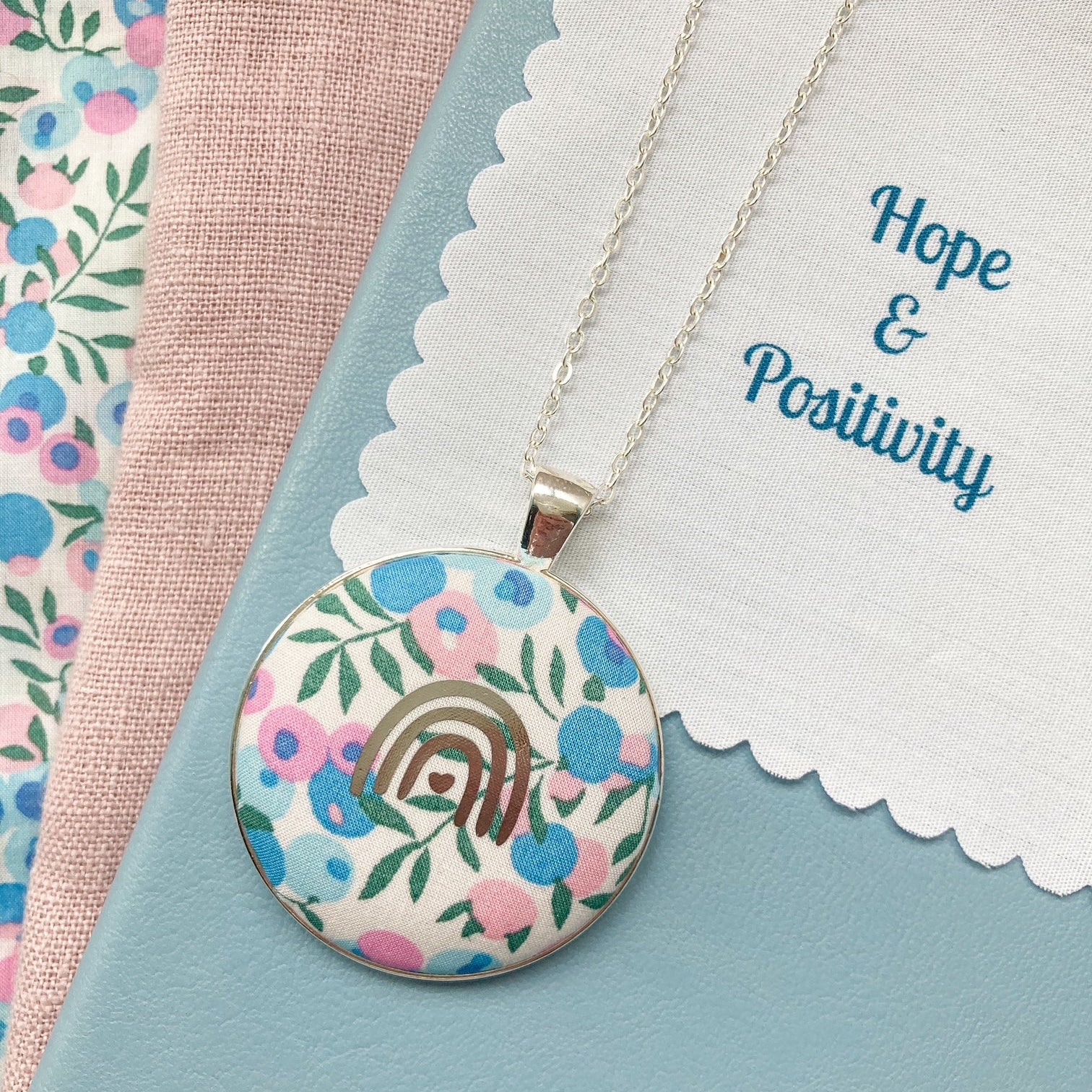Necklace with floral pendant with a fabric message saying 'Hope & Positivity', against a floral and pink fabric background.