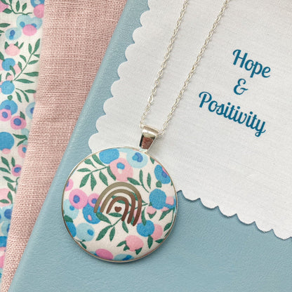 Necklace with floral pendant with a fabric message saying 'Hope & Positivity', against a floral and pink fabric background.