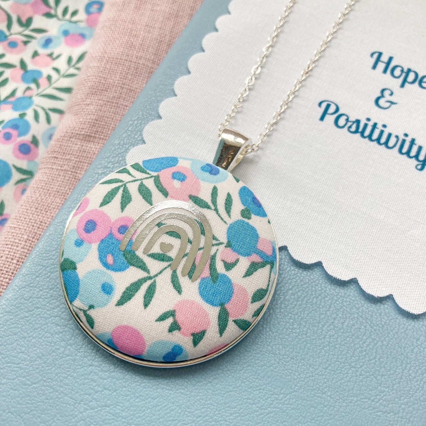 Floral pendant necklace on a blue background with a fabric message that says 'Hope & Positivity'.