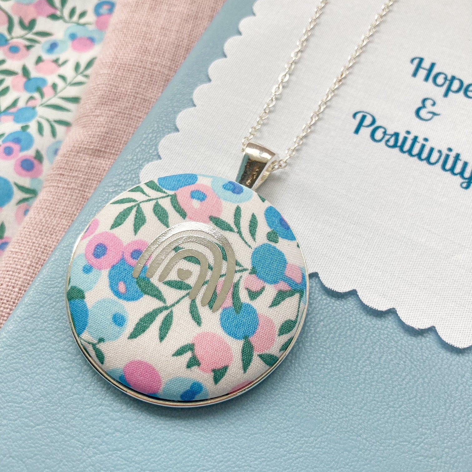 Floral pendant necklace on a blue background with a fabric message that says 'Hope & Positivity'.