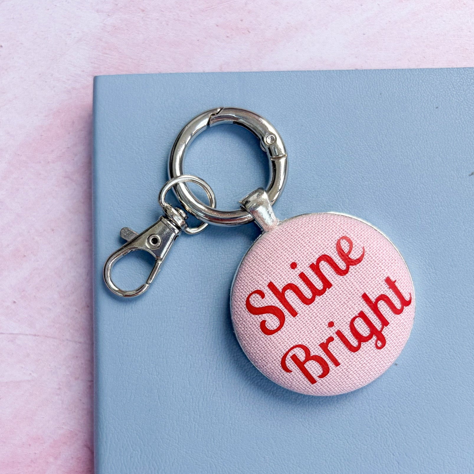 Keyring with 'Shine Bright' in red on pink fabric, set on a pink background