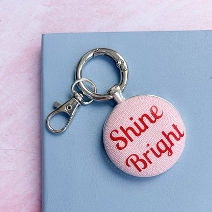 Keyring with 'Shine Bright' in red on pink fabric, set on a pink background