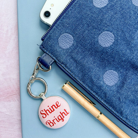 Blue polka dot pouch with a "Shine Bright" red and pink keyring on a light pink background