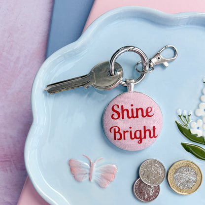 Keyring with 'Shine Bright' words in red on pink fabric, on a decorative plate with coins and keys, on a pink and blue background.