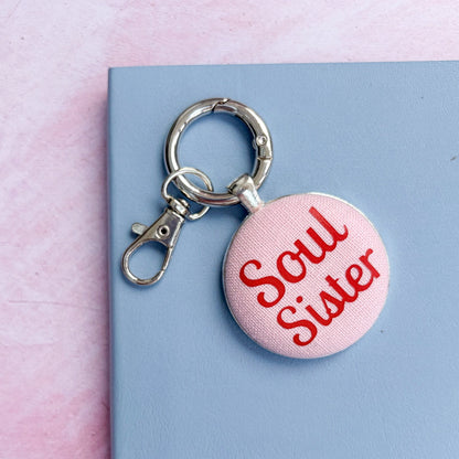 Keyring with 'Soul Sister' printed in red on pink fabric, set on a blue notebook and pink background