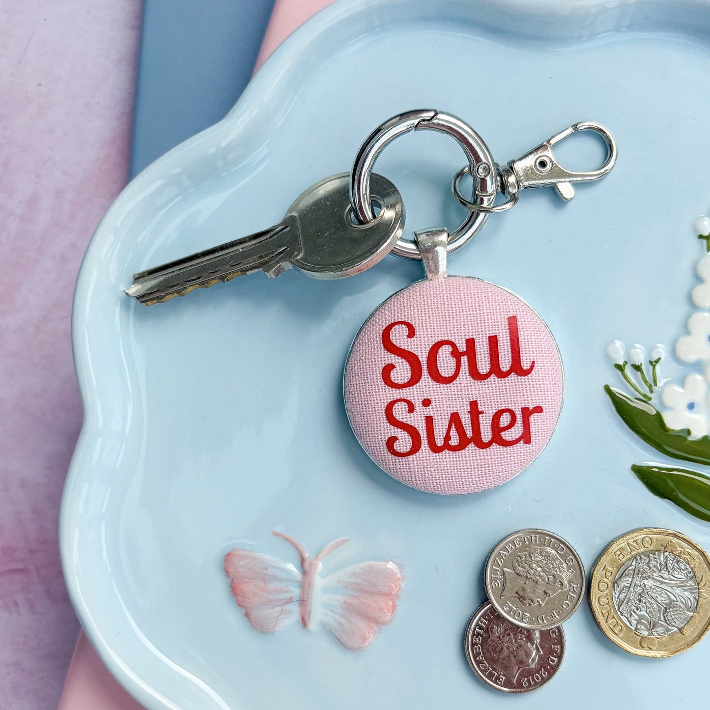 Keyring with 'Soul Sister' printed in red on pink fabric, on a decorative plate with coins and flowers