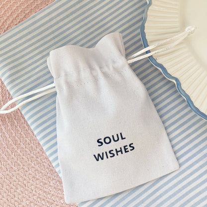 White drawstring gift pouch with 'Soul Wishes' text, on a blue and white striped fabric background