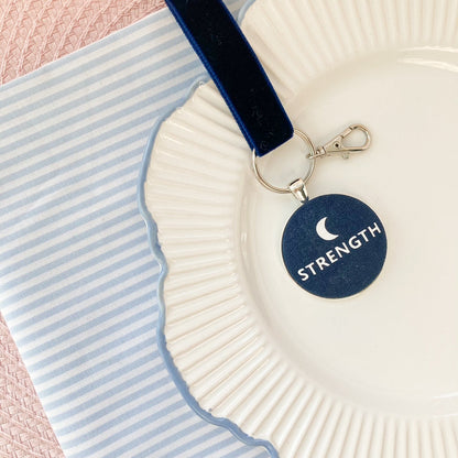 Keychain with 'STRENGTH' and a new moon on a decorative plate with striped background