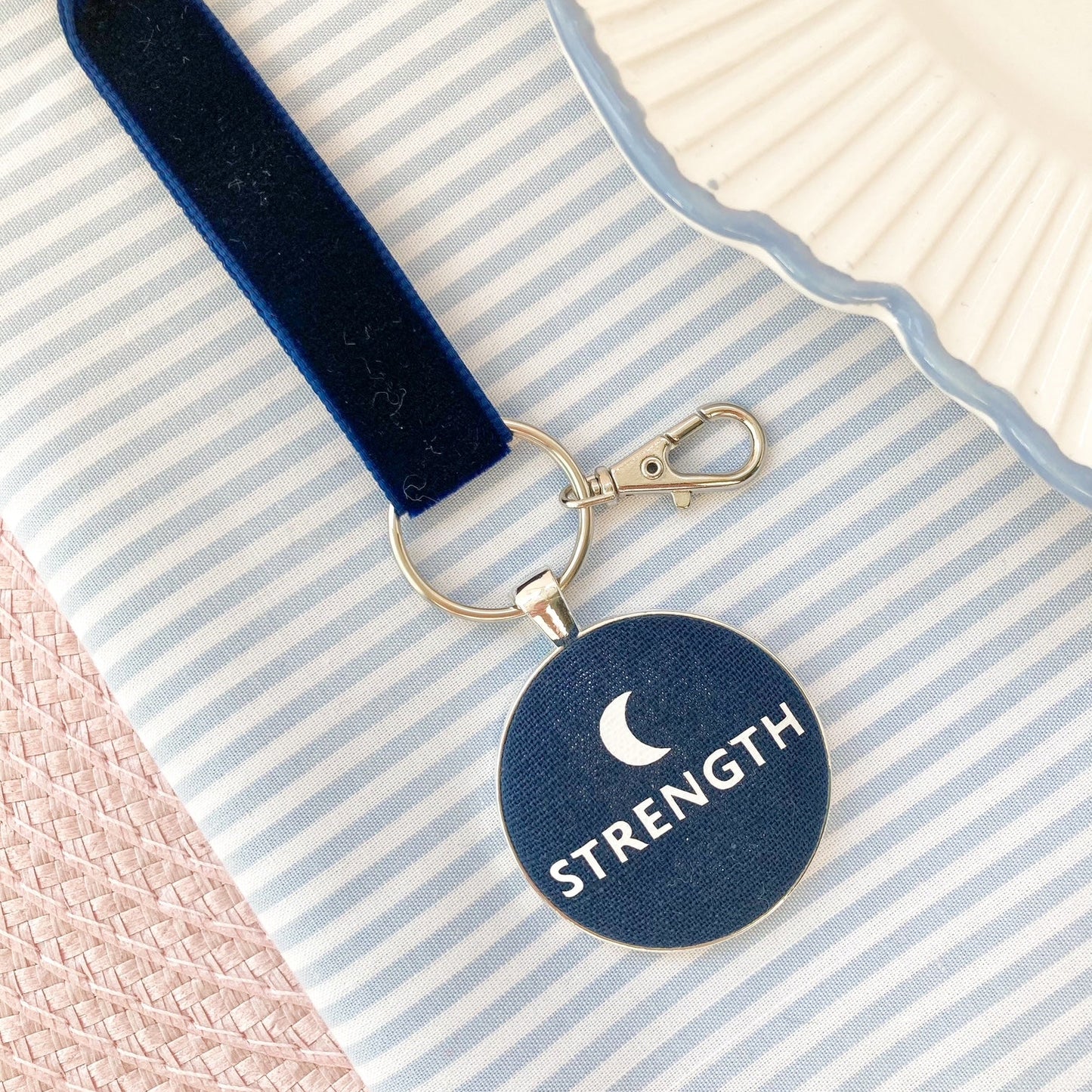 Navy blue key ring with 'STRENGTH' and moon symbol on a striped fabric background