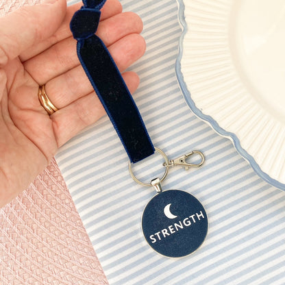 Navy blue keepsake with 'STRENGTH' and a new moon design held by a hand on a striped background