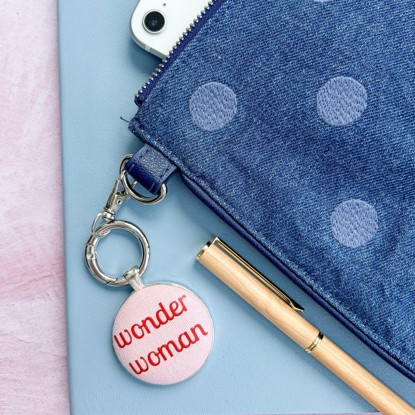 Pink 'wonder woman' bag charm on a blue polka dot pouch with a phone and pen.