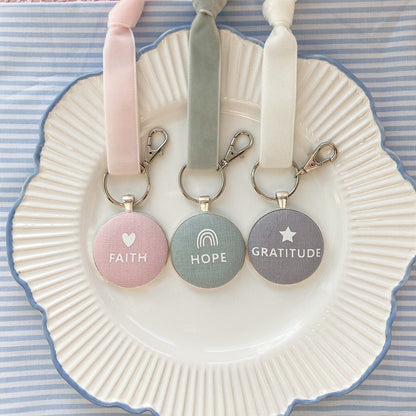 Three key rings and keepsakes with 'Faith', 'Hope', and 'Gratitude' printed on fabric, laid on a decorative plate.