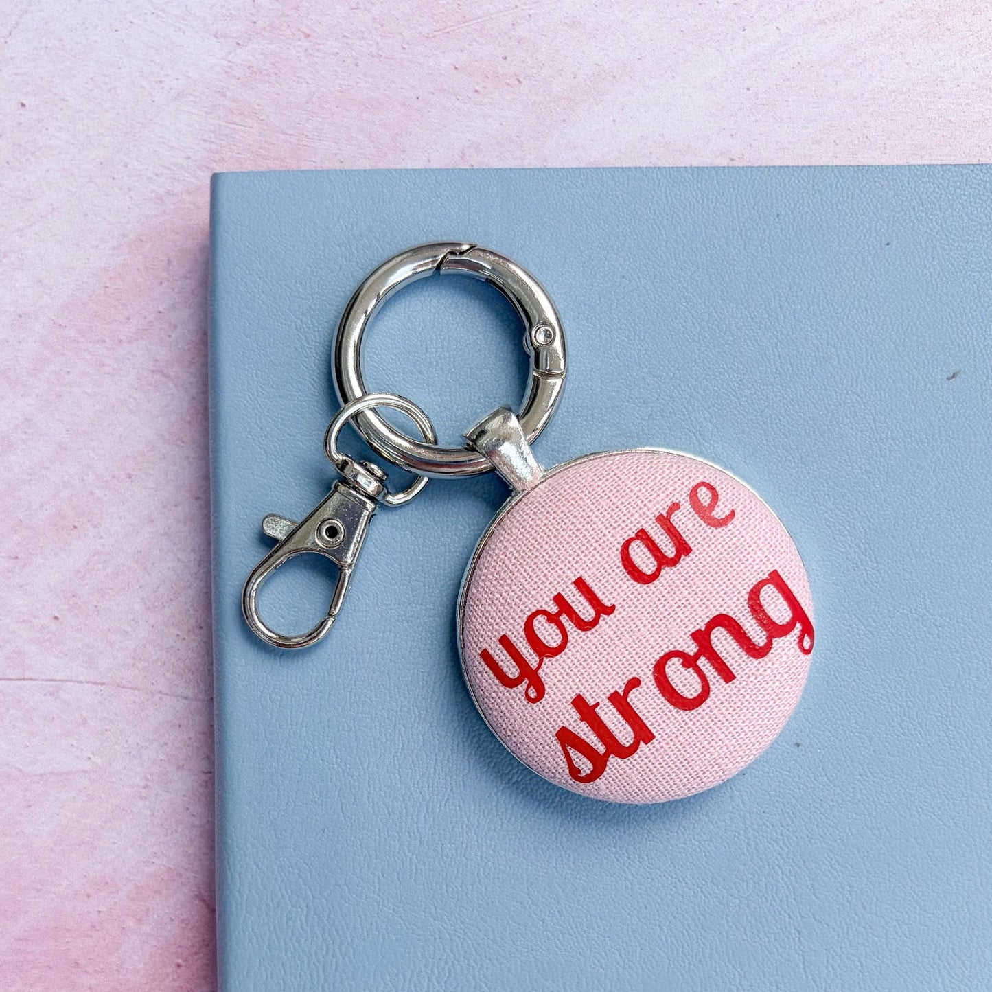Keyring with 'you are strong' red words printed on pink fabric, set on a blue notebook and pink background