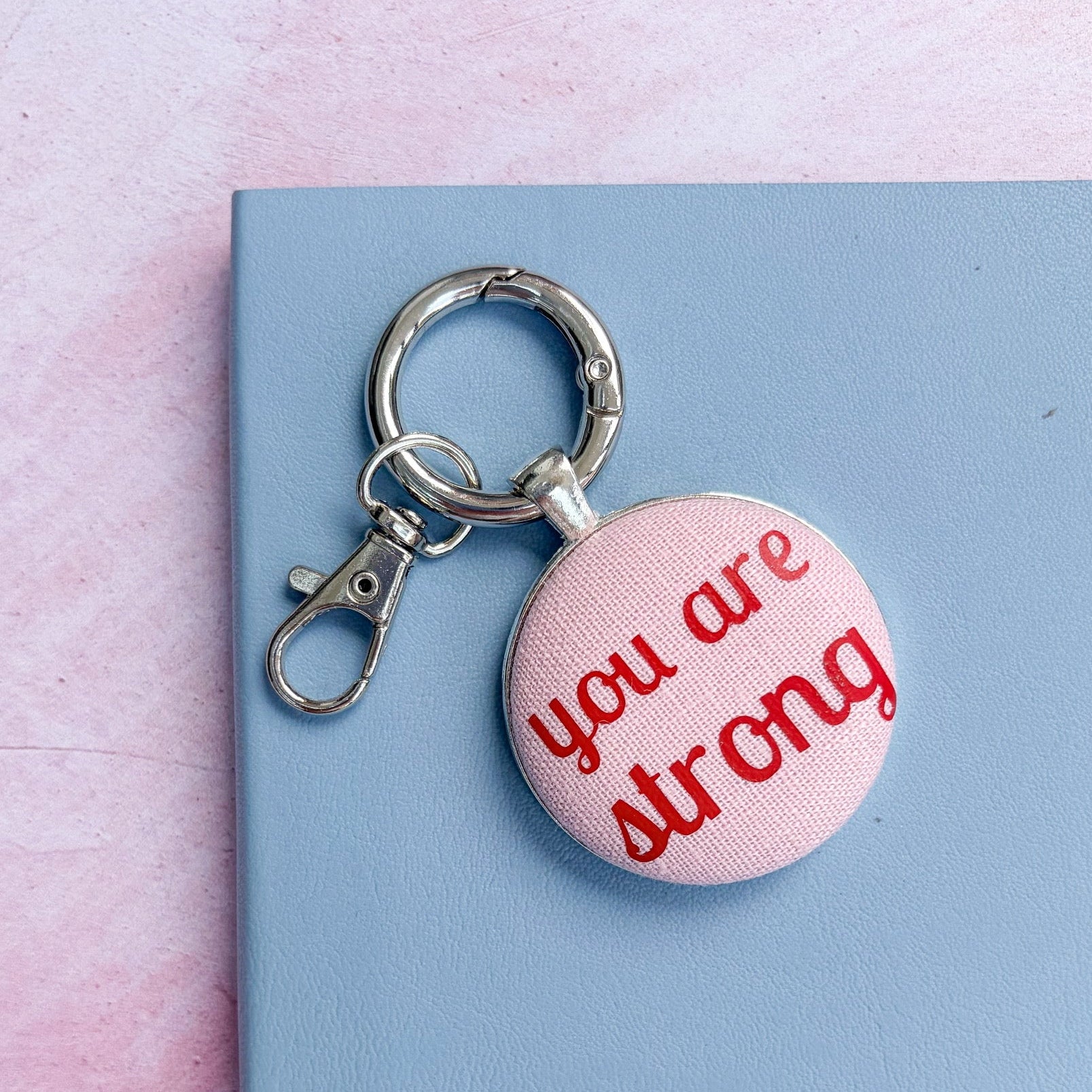 Keyring with 'you are strong' red words printed on pink fabric, set on a blue notebook and pink background