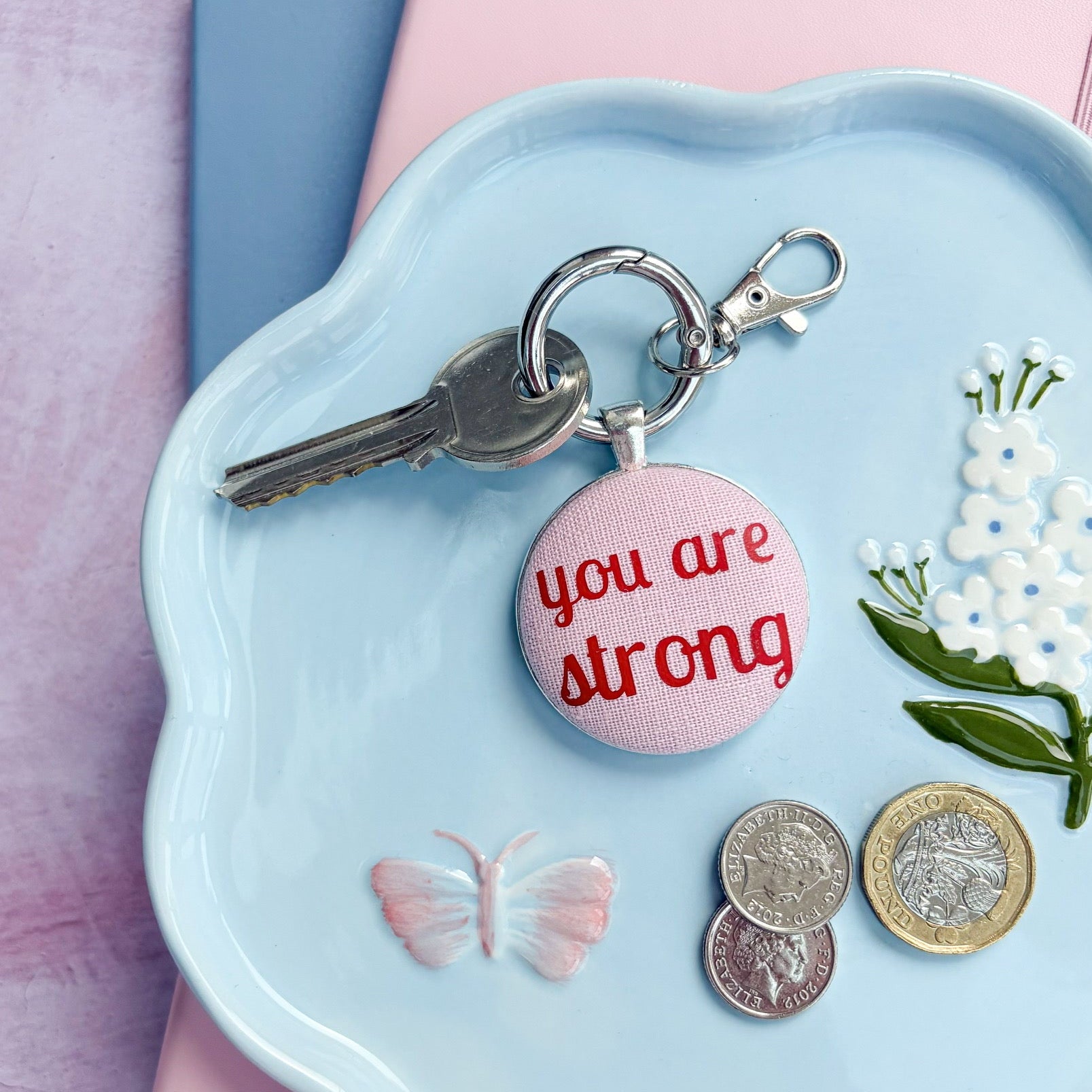 Light blue dish with a 'you are strong' keyring in pink and red, coins, and flowers on a pink background