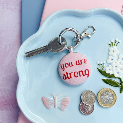 Light blue dish with a 'you are strong' keyring in pink and red, coins, and flowers on a pink background