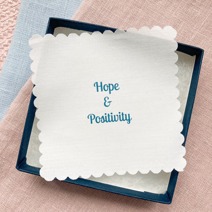 White fabric message with 'Hope & Positivity' text in a blue box on a pink and blue fabric background