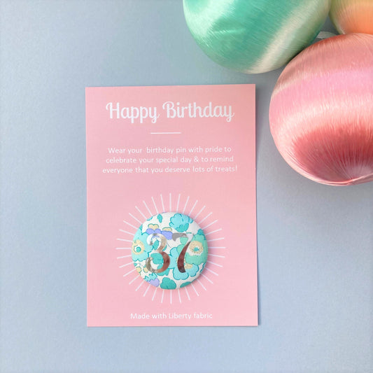 A Happy Birthday fabric pin with a floral pattern in mint green and printed silver foil, featuring an age number '37' in the centre, presented on a pink happy birthday postcard.
