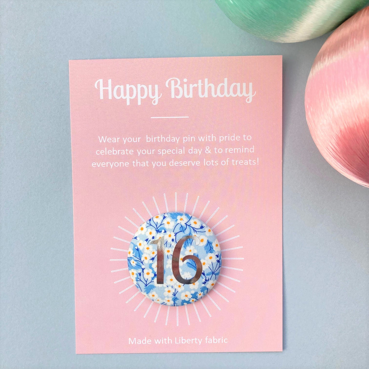 A round birthday pin with blue floral pattern and the number 16 in silver foil, indicating it is suitable for a 16th birthday, attached to a birthday postcard.