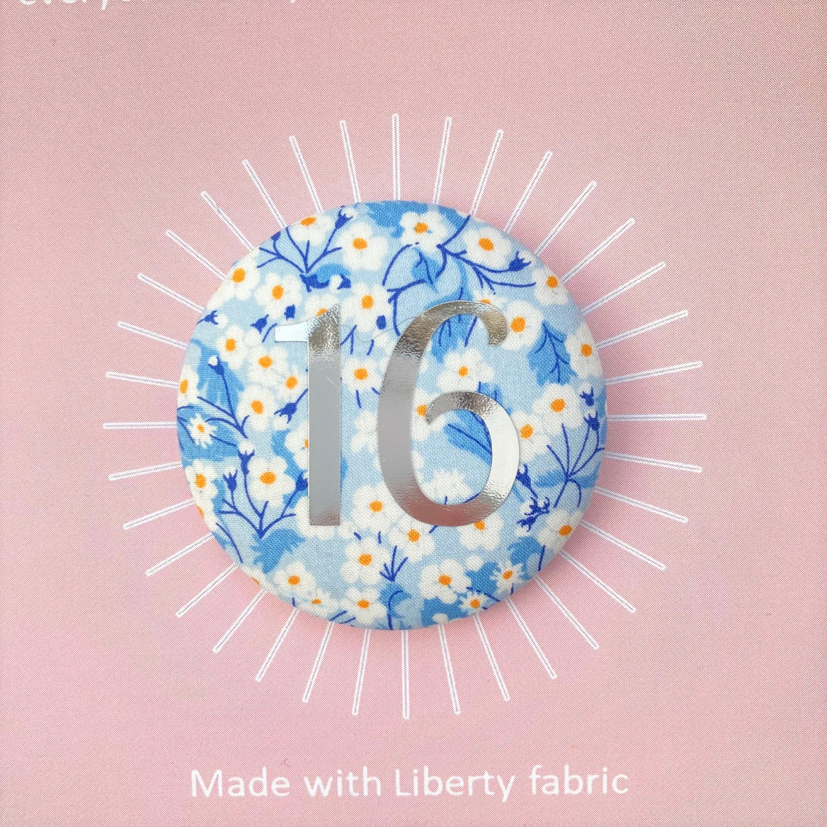A round birthday pin with blue floral pattern and the number 16 in silver foil, indicating it is suitable for a 16th birthday.
