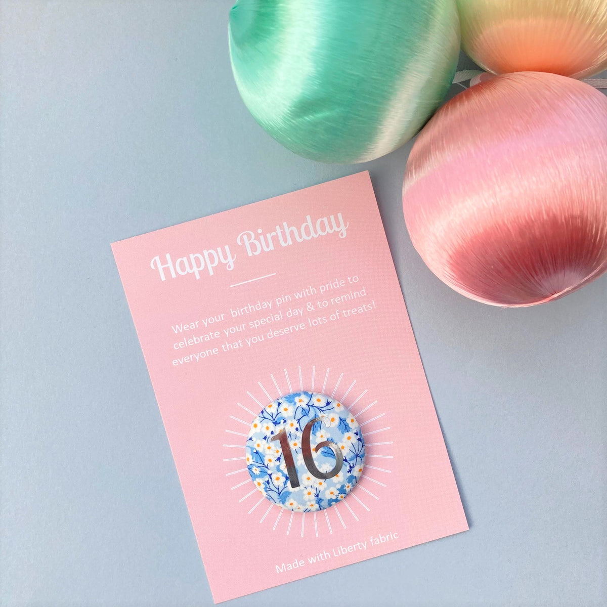 A round birthday pin with blue floral pattern and the number 16 in silver foil, indicating it is suitable for a 16th birthday.