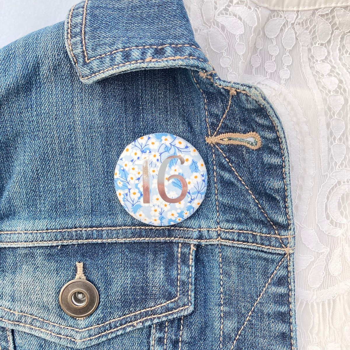 A round birthday pin with blue floral pattern and the number 16 in silver foil, indicating it is suitable for a 16th birthday.