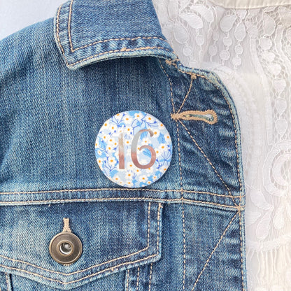 A round birthday pin with blue floral pattern and the number 16 in silver foil, indicating it is suitable for a 16th birthday.