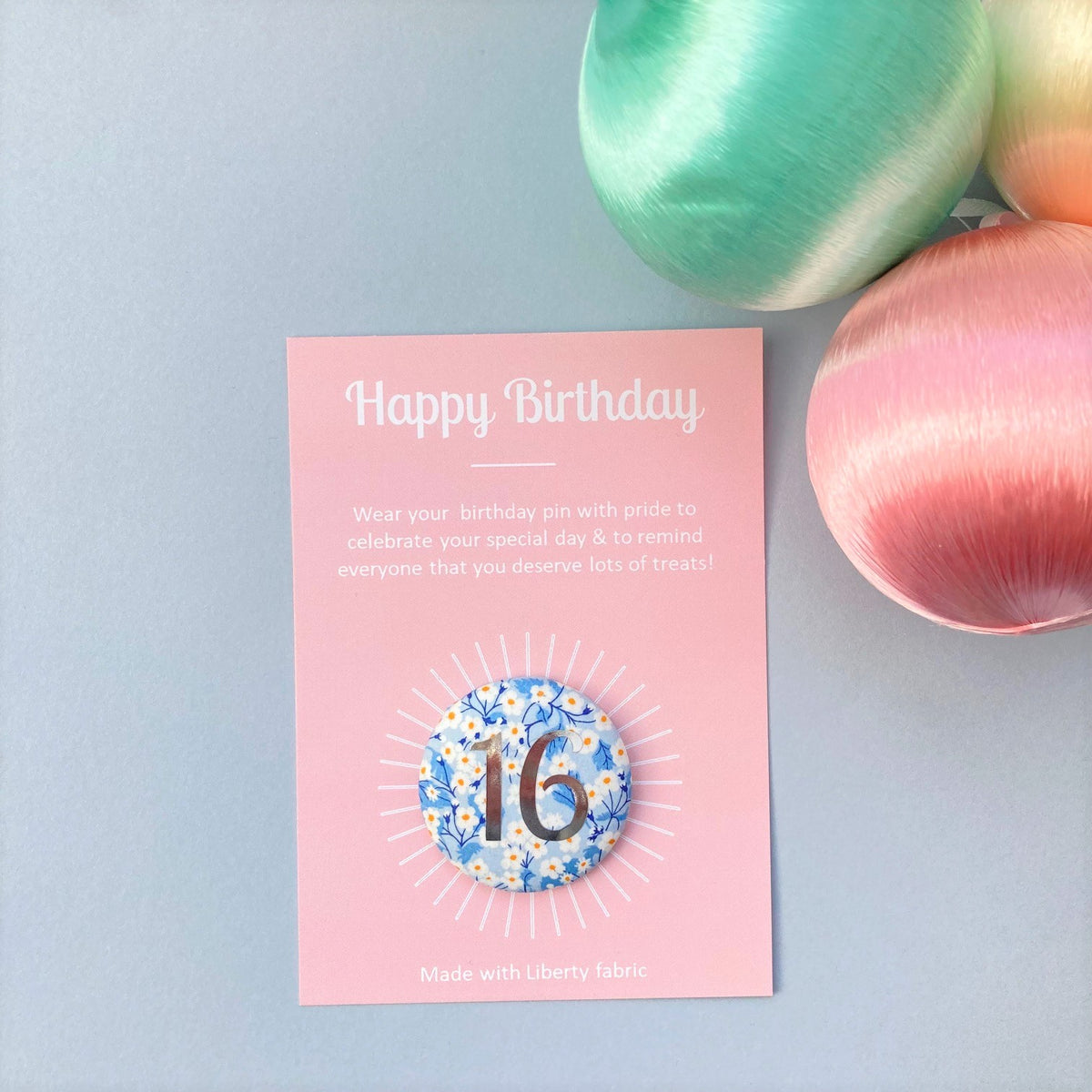 A round birthday pin with blue floral pattern and the number 16 in silver foil, indicating it is suitable for a 16th birthday, attached to a birthday postcard.