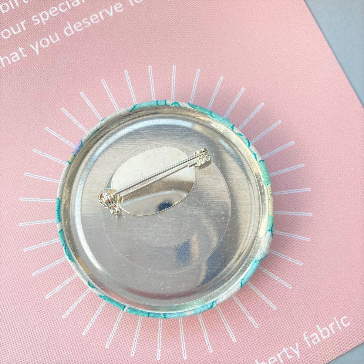 The reverse and pin of a round birthday pin with blue floral pattern and the number 16 in silver foil, indicating it is suitable for a 16th birthday, attached to a birthday postcard.