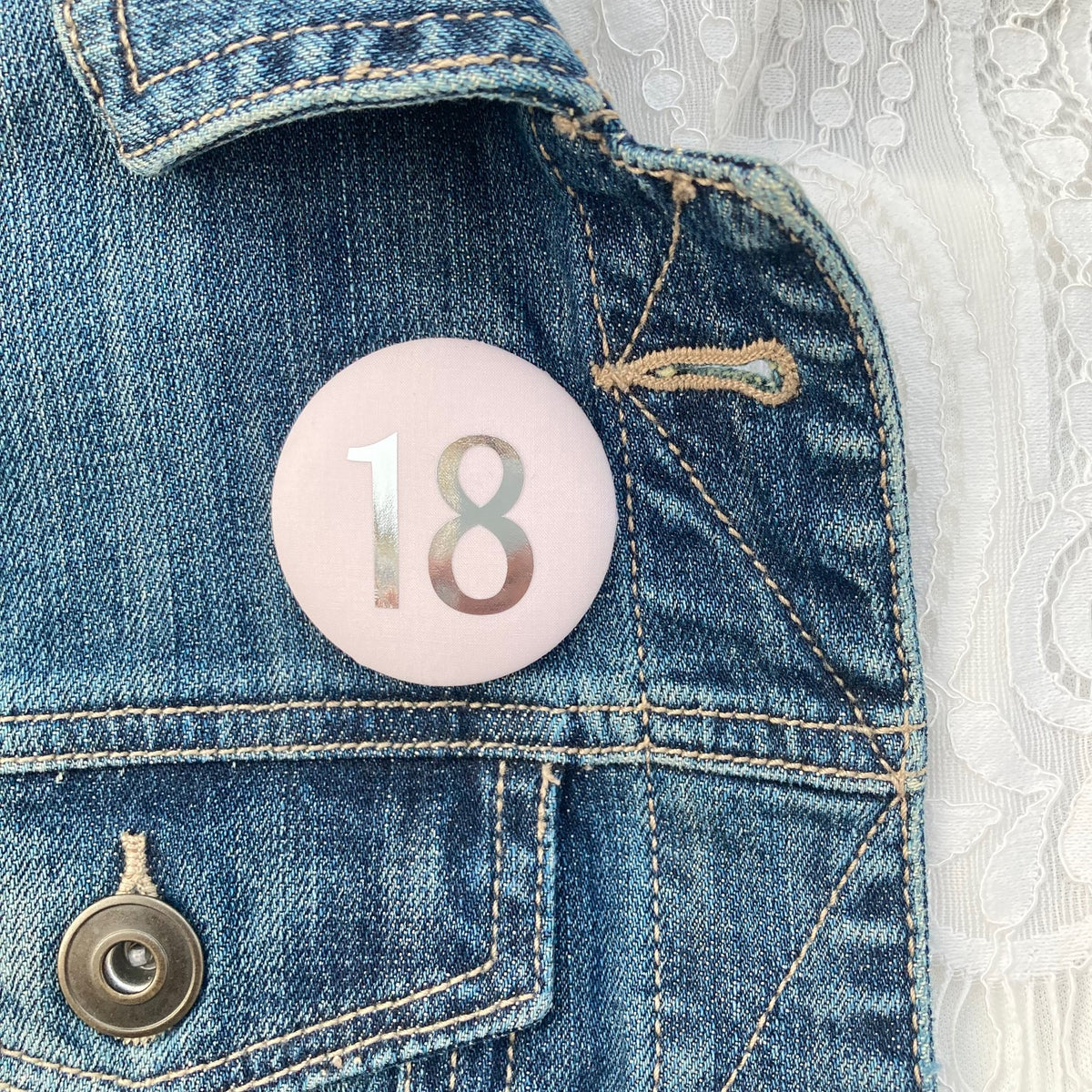A fabric pale pink '18' birthday pin with a silver plated brooch fastening, attached to a denim jacket.