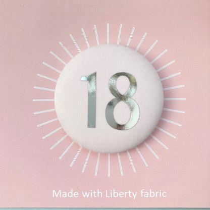 A fabric pale pink '18' birthday pin with a silver plated brooch fastening, attached to a birthday postcard