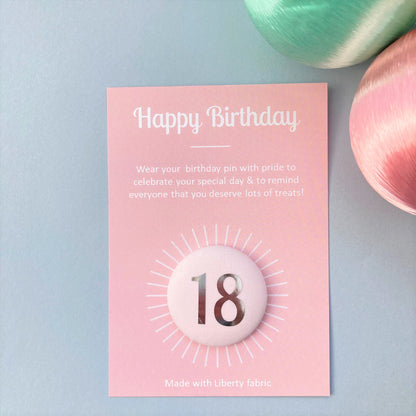 A fabric pale pink '18' birthday pin with a silver plated brooch fastening, attached to a birthday postcard